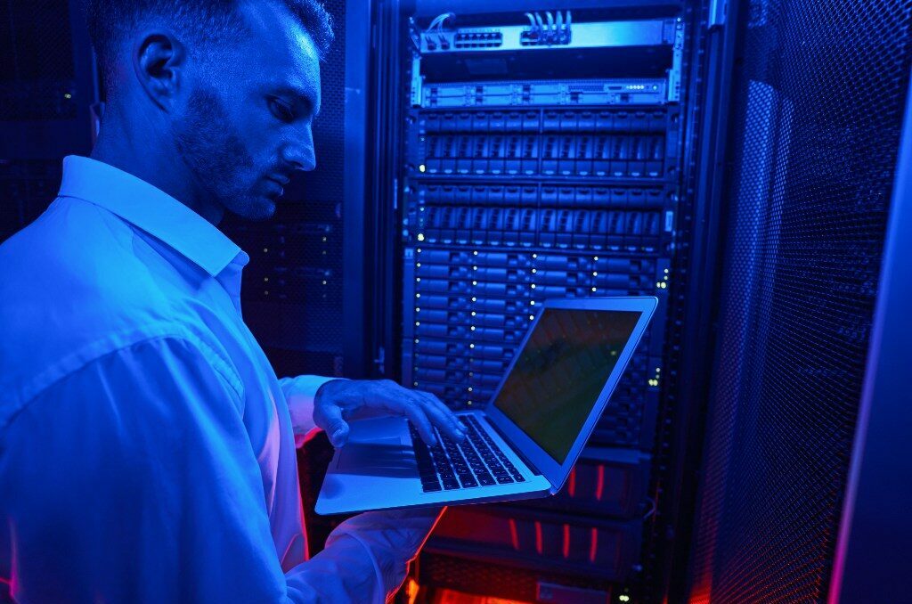 man in blue-lit server room holds laptop